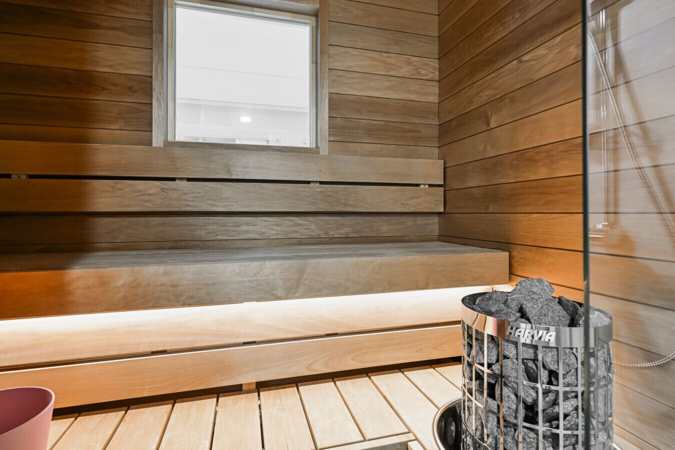 A sauna with wooden benches, a window, and a metal container with rocks.
