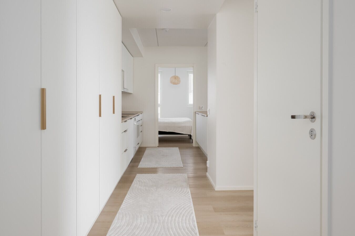 A narrow hallway with white walls, wooden flooring, and a bed at the end.