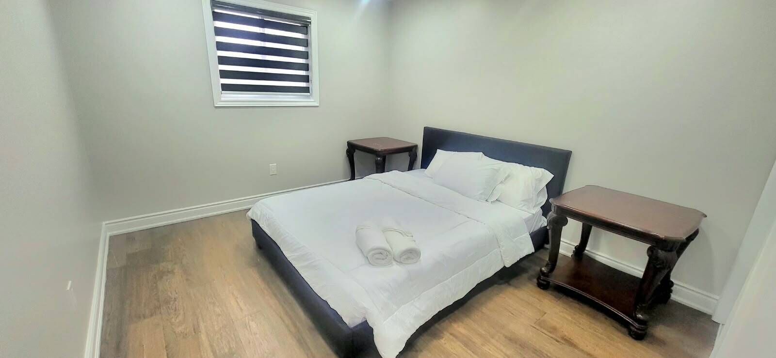A bedroom with a bed, two nightstands, a window with blinds, and a wooden floor.