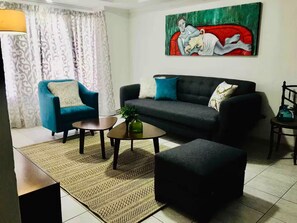 Ein Wohnzimmer mit einem blauen Sessel, einem grauen Sofa, einem kleinen Couchtisch und einem schwarzen Hocker. Über dem Sofa hängt ein Gemälde.