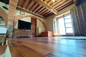 A spacious living room with a wooden floor, a television on a wooden cabinet, and a large window with curtains. - Grandma Isola's House (Prata)