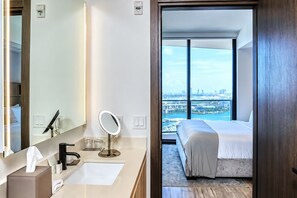 A hotel room with a large mirror, a sink, a bedside table, and a bed. There is a view of a cityscape through large windows. - Stylish Ocean-View Condo in Downtown Miami (Miami)