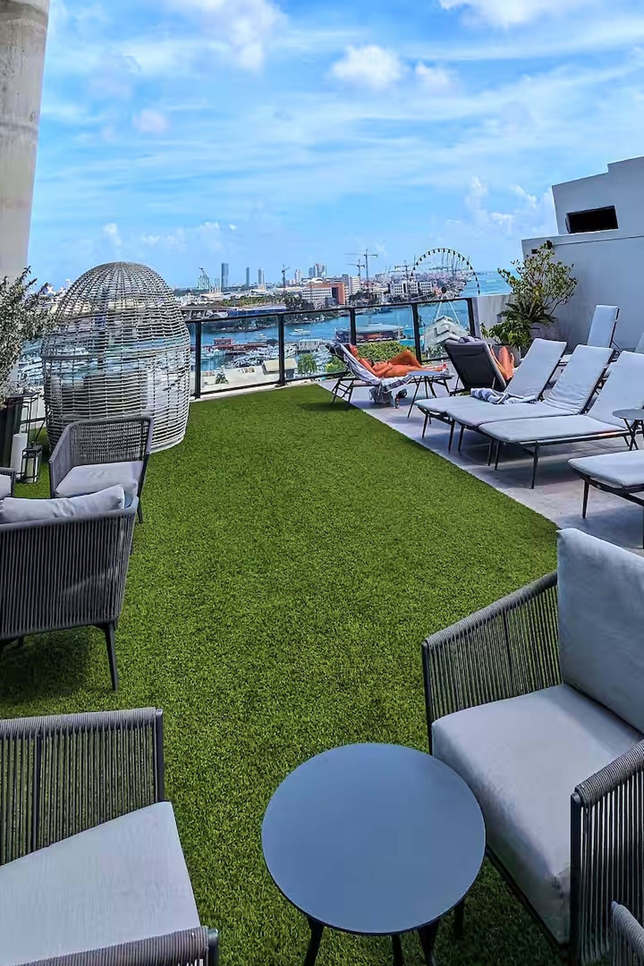 Terraço no telhado com grama sintética, cadeiras de descanso e vista para um porto e horizonte urbano.