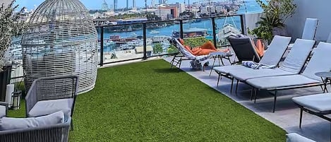 A rooftop terrace with artificial grass, lounge chairs, and a view of a marina and city skyline.