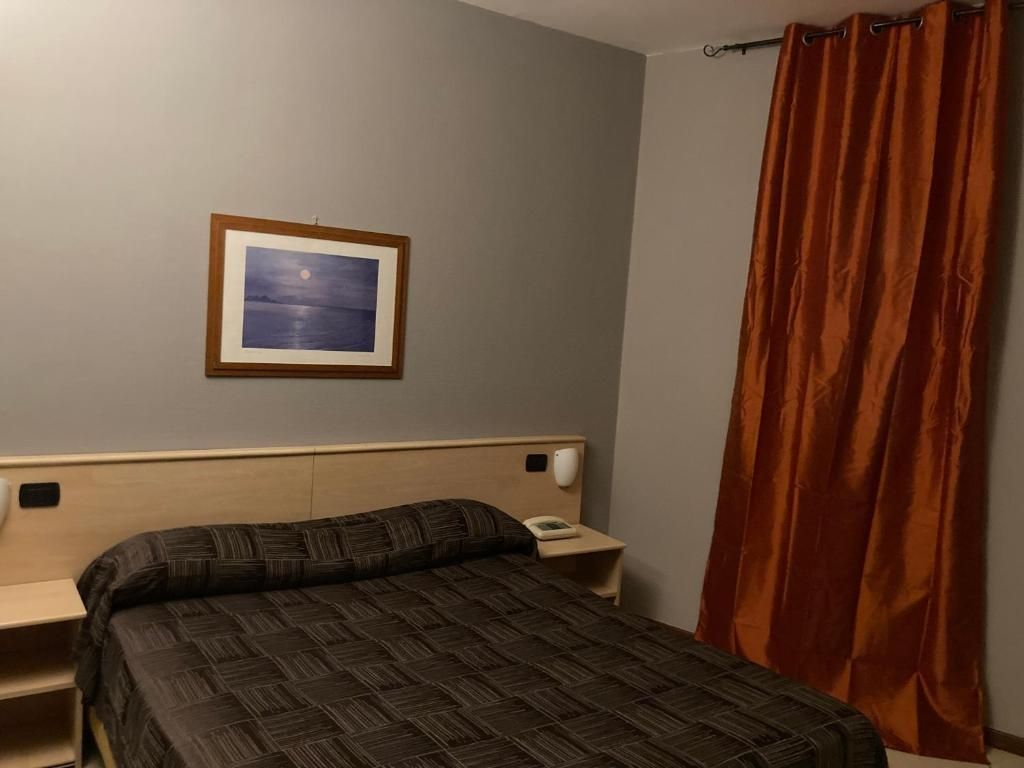 Ein Hotelzimmer mit einem Bett, einem Nachttisch, einem Telefon, einem gerahmten Bild an der Wand und einem Vorhang.