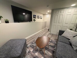 Ein modernes Wohnzimmer mit einem an der Wand befestigten Flachbildfernseher, einem grauen Sofa mit gemusterten Kissen, einem kleinen Holztisch und einem Teppich mit Muster.
