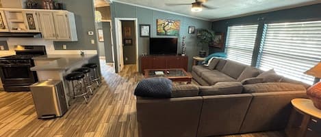 Una sala de estar moderna con un sofá seccional, una mesa de centro y un televisor. Hay una zona de cocina con un refrigerador, estufa y taburetes de bar.