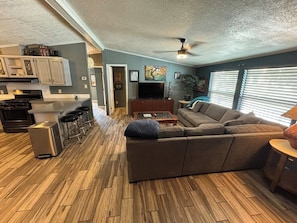 A modern living room with a sectional sofa, a coffee table, and a TV. There is a kitchen area with a refrigerator, stove, and bar stools.