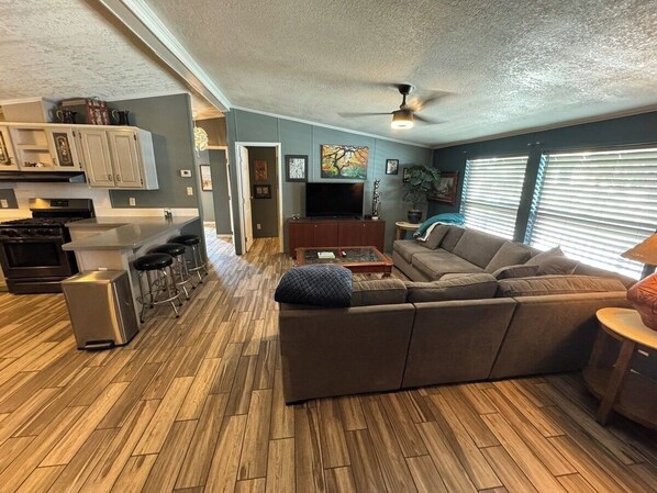 A modern living room with a sectional sofa, a coffee table, and a TV. There is a kitchen area with a refrigerator, stove, and bar stools.