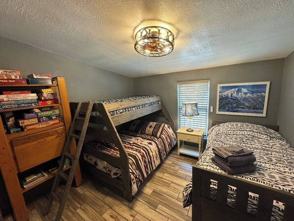 A room with a bunk bed, a ladder, a bookshelf with board games, a bedside table with a lamp, and a framed picture on the wall.