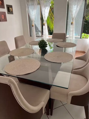 A modern dining area with a glass table surrounded by beige chairs, a small potted plant, and a view of greenery outside. - Resort 5 Estrellas - Vista a Piscina & Jacuzzi (Santa Cruz de la Sierra)