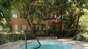 A stone hot tub with steps leading into it, surrounded by greenery and a building in the background.