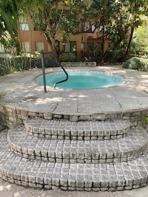 A stone hot tub with steps leading into it, surrounded by greenery and a building in the background.