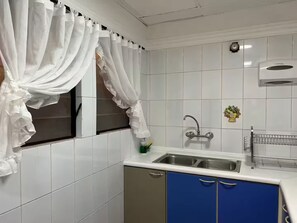 Een keuken met witte betegelde muren, een roestvrijstalen spoelbak, blauwe kastjes en een raam met witte gordijnen.