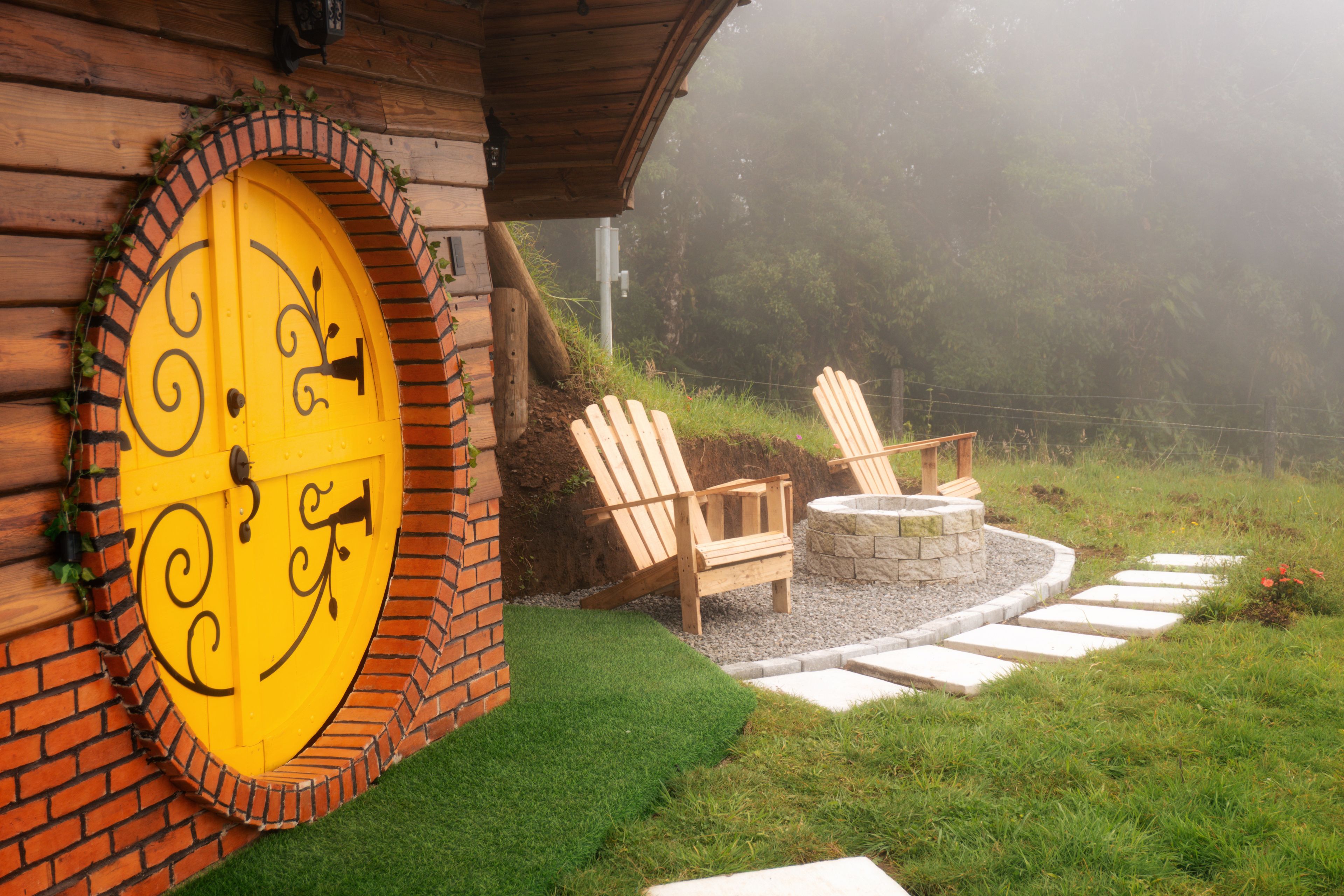 A wooden house with a yellow door, a brick wall, and a garden with a fire pit and Adirondack chairs.