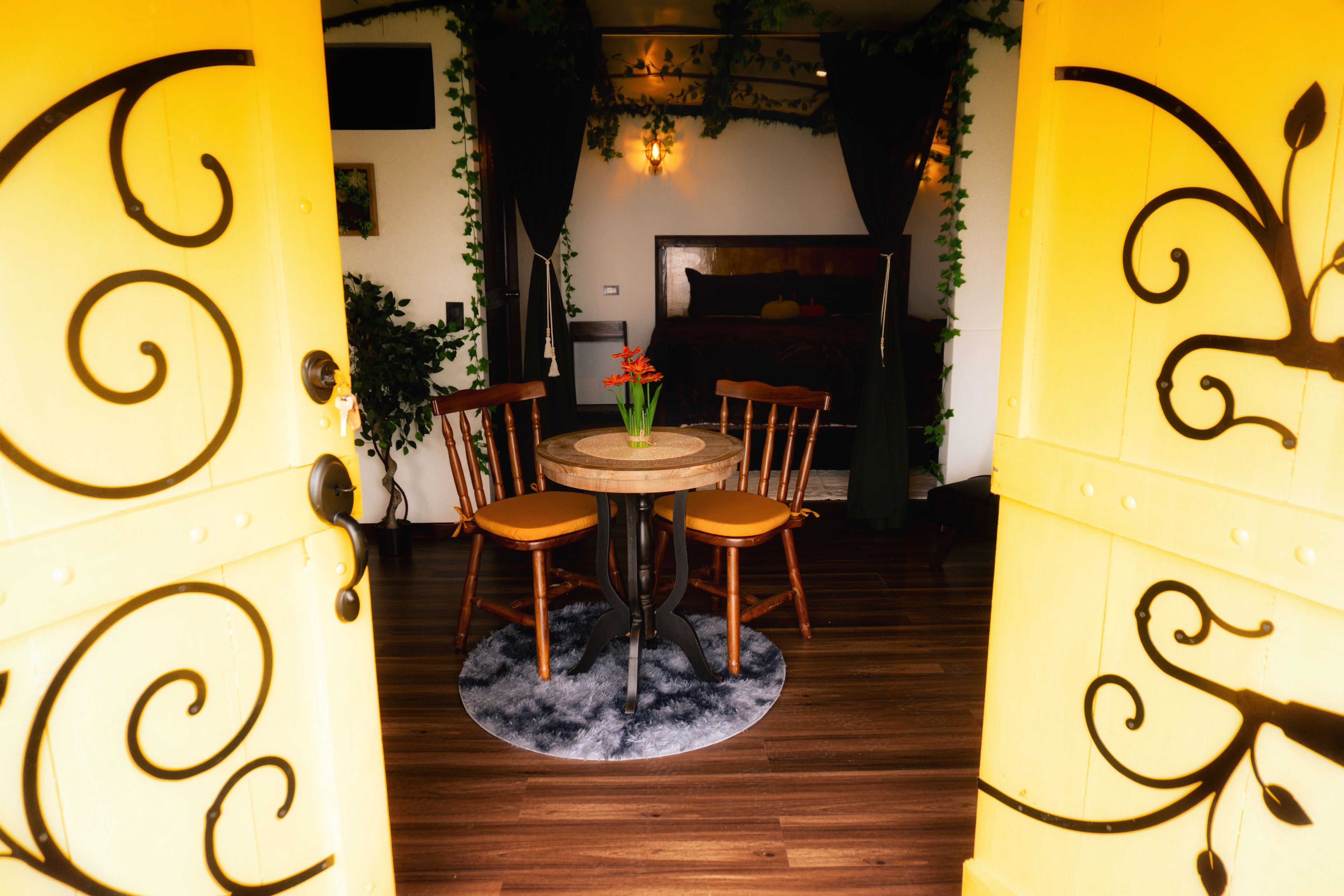 A room with a round table, two chairs, a small vase with flowers, and a door with decorative ironwork.