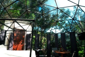 Estructura de domo geodésico con una cama, una mesa y sillas en su interior, rodeada de exuberante vegetación.