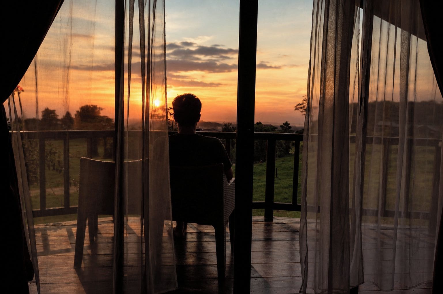 A person standing on a wooden deck with a view of the sunset through sheer curtains.