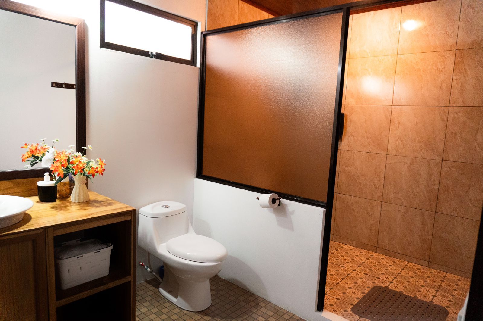 Une salle de bain avec des toilettes, un lavabo et un miroir. On y trouve une étagère avec un récipient et un vase avec des fleurs.