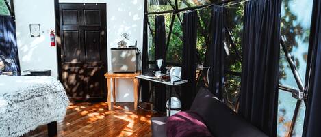 Una habitación con suelo de madera, un sofá con un cojín, una mesita con una tetera, un televisor y una puerta.