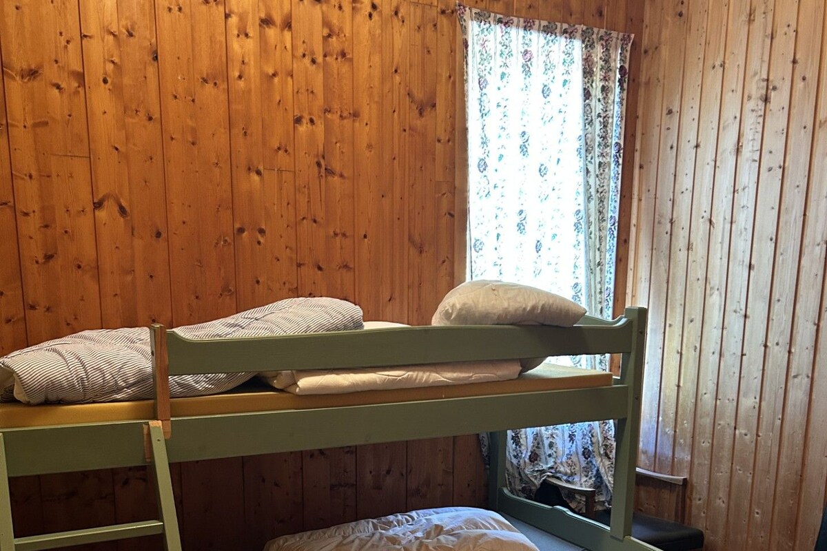 A bunk bed room with wooden walls, a window with floral curtains, and a small table.