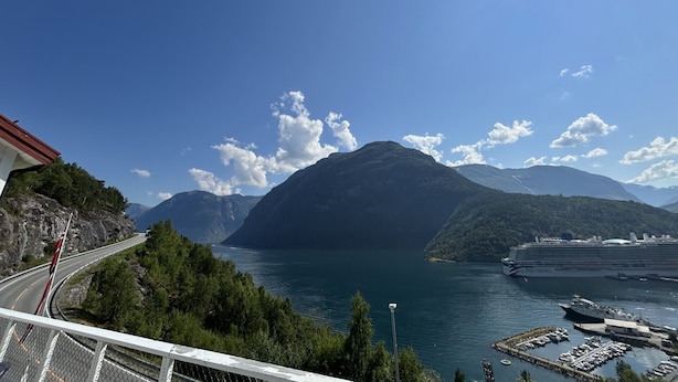 Un paisaje costero con una carretera, montañas y un crucero amarrado en un puerto deportivo.