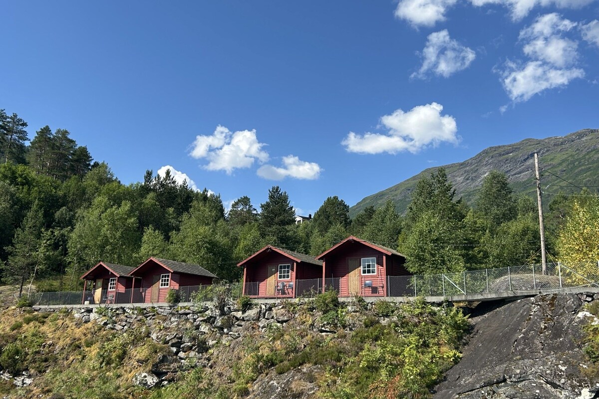 Three red cabins with a stone wall in front, set against a backdrop of trees and a mountain.
