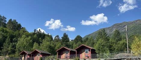 Three red cabins with a stone wall and a metal fence in front, set against a backdrop of trees and a mountain.