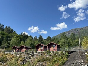 Tres cabañas rojas con un muro de piedra y valla de metal, frente a un fondo de árboles y una montaña.