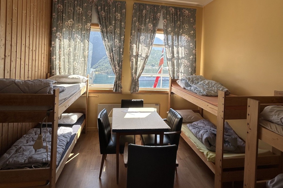 A room with bunk beds, a table, and chairs, with a view of the sea and mountains through the window.