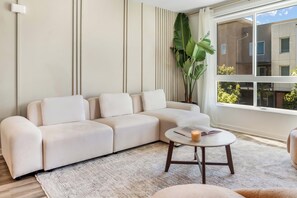 A modern living room with a sectional sofa, a round coffee table, and a large window offering a view of buildings outside. - Casa Monica® Luxury 2BR in Irvine w/ Gym, Pool (Irvine)