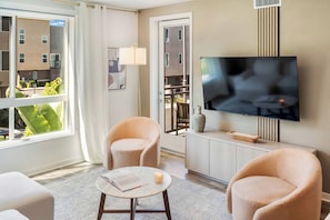 A modern living room with a flat-screen TV, two armchairs, a small round table with a book and candle, and a view of the outside through large windows.