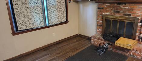 A room with a fireplace, a wooden floor, a patterned window, and a brick wall.