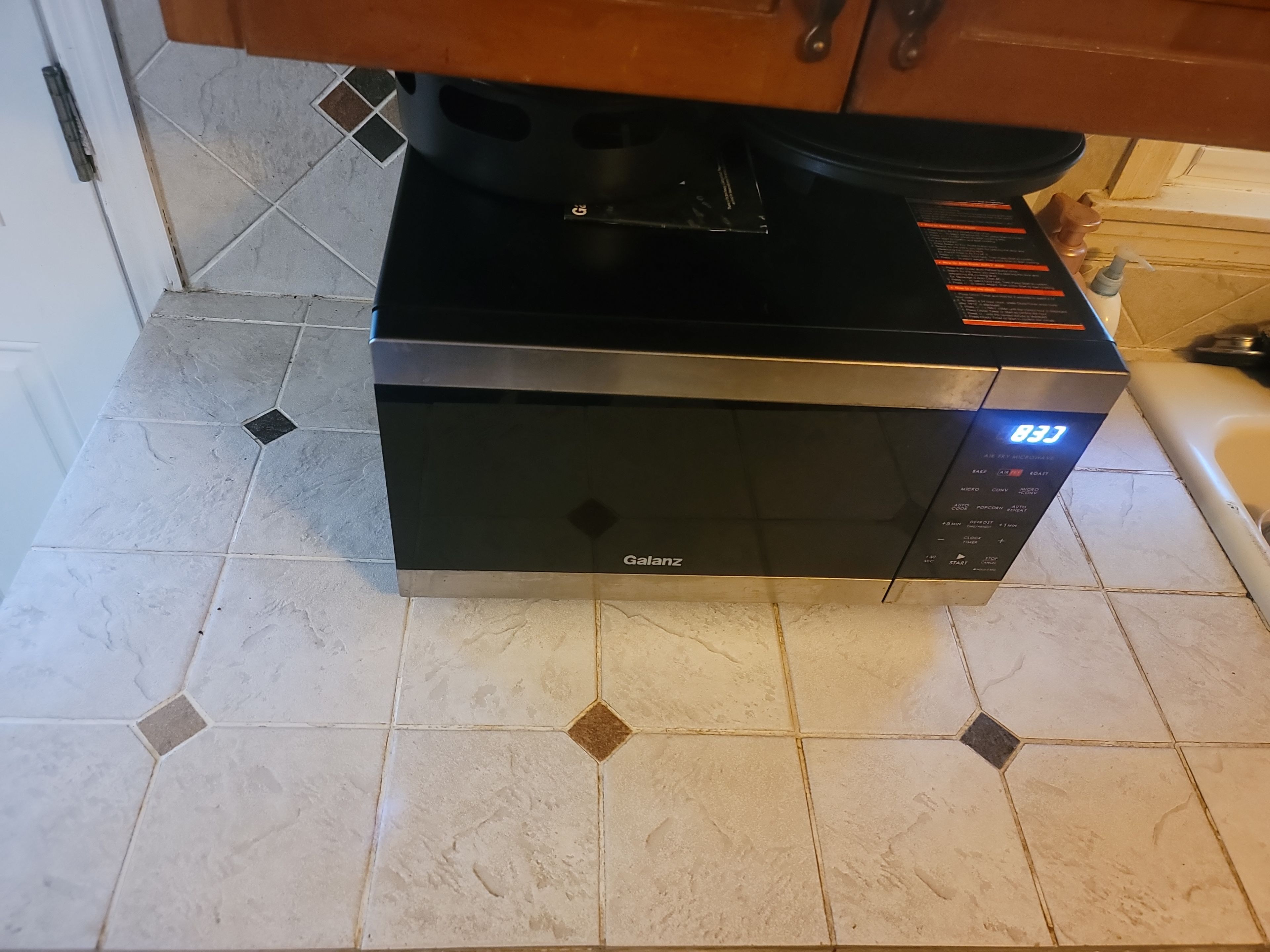 Un horno microondas negro y plateado con pantalla digital, sobre un piso de baldosas con diseño de rombos.
