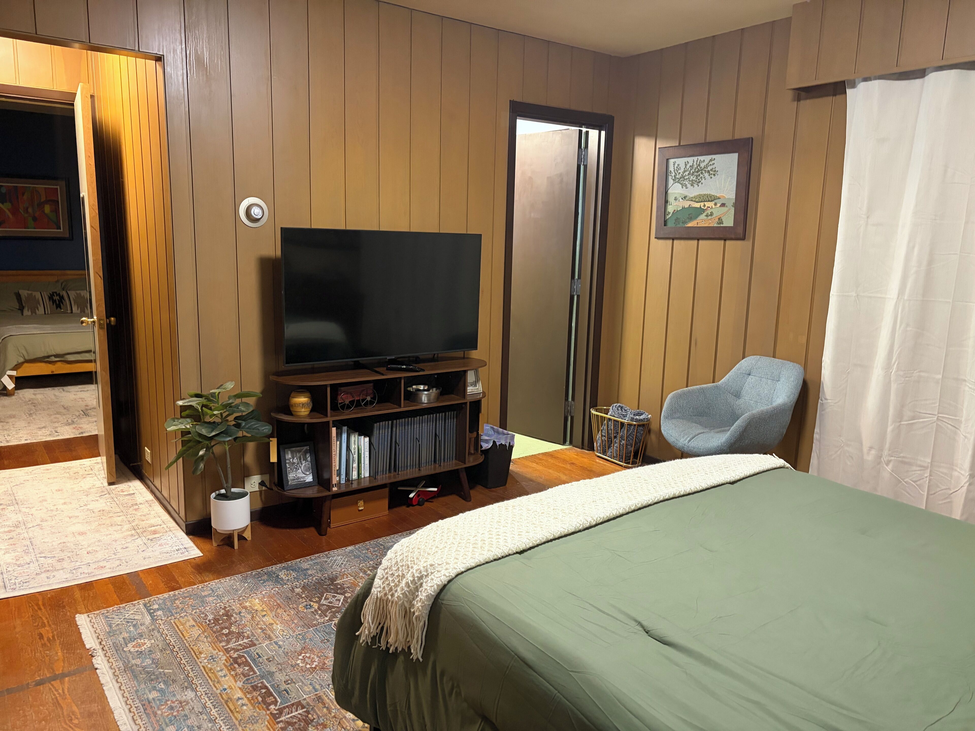 A bedroom with a bed, a television on a wooden stand, a chair, and a door leading to another room.