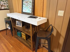 A small, wooden-framed desk with a white marble top, a black notebook, and keys. Below the desk, there is a shelf with a framed picture and a small plant. - The Thoroughbred Private 2-Bedroom Historic Suite (Prineville)