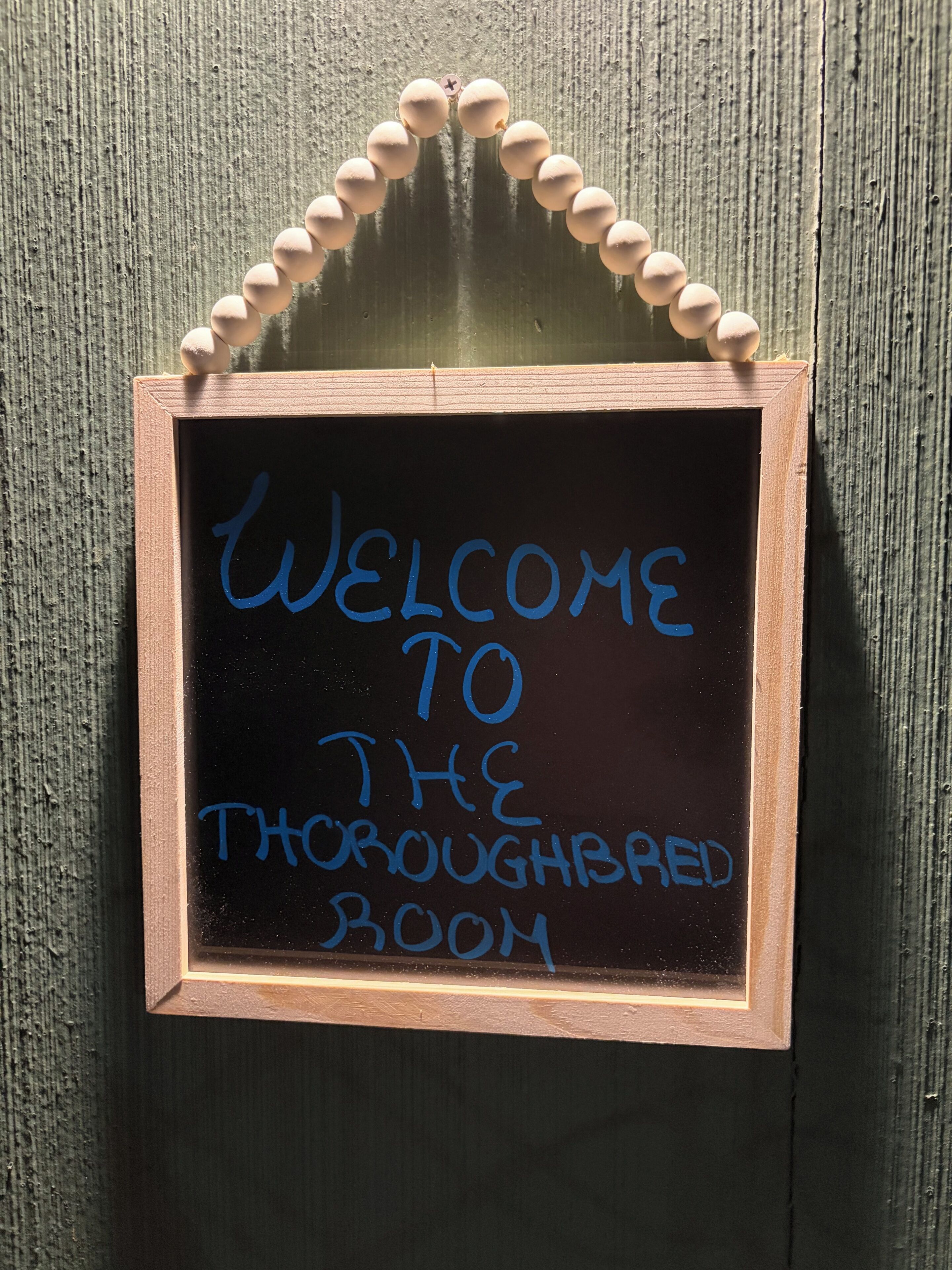 A chalkboard sign with the message 'WELCOME TO THE THOROUGHBRD ROOM' mounted on a wooden wall.