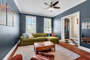 Ein Zimmer mit einem grünen Sofa, einem Holztisch und einem braunen Ledersessel. Es gibt zwei Fenster mit Jalousien, einen Deckenventilator und einen Schachbrettfußboden.