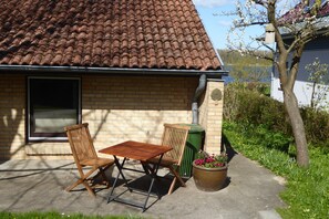 A house with a tiled roof, a brick wall, and a window. There is a green trash bin and a flower pot with flowers. - "Am Binnenwasser" Vacation Rental: Apartment with an alcove bedroom, kitchen, and large sunroom (Neustadt in Holstein)