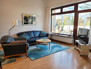 A living room with a sofa, a coffee table, a TV on a stand, and a large window. - "Am Binnenwasser" Vacation Rental: Apartment with an alcove bedroom, kitchen, and large sunroom (Neustadt in Holstein)