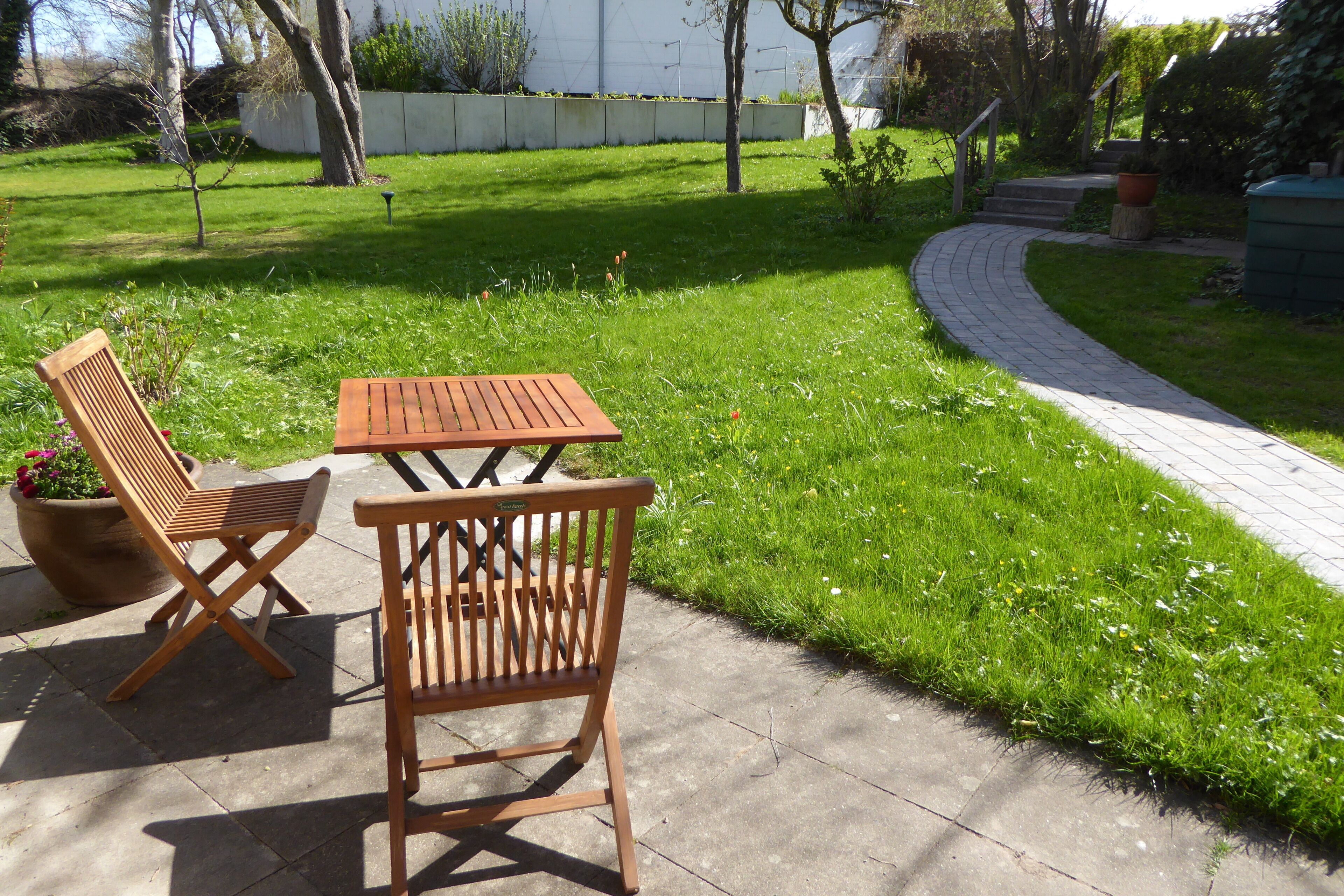 Eine Terrasse mit zwei Holzstühlen und einem Tisch, umgeben von einem gepflegten Rasen und einem gepflasterten Weg.