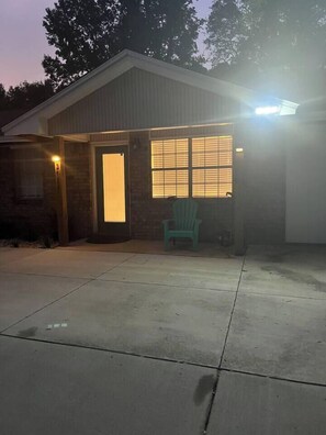 Un bâtiment d’un seul étage, doté d’une porte de garage, d’un petit porche avec une chaise et d’une fenêtre éclairée.