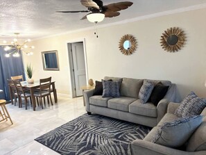 Un salon avec un canapé gris, un tapis à motifs, un ventilateur de plafond, des éléments de décoration murale et un coin repas avec des meubles en bois.