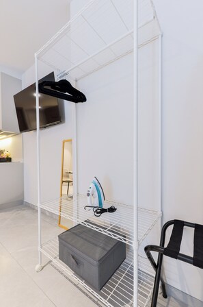 Een kleine kamer met een wit metalen schap, een grijze opbergdoos, een zwarte stoel en een aan de muur bevestigde televisie.