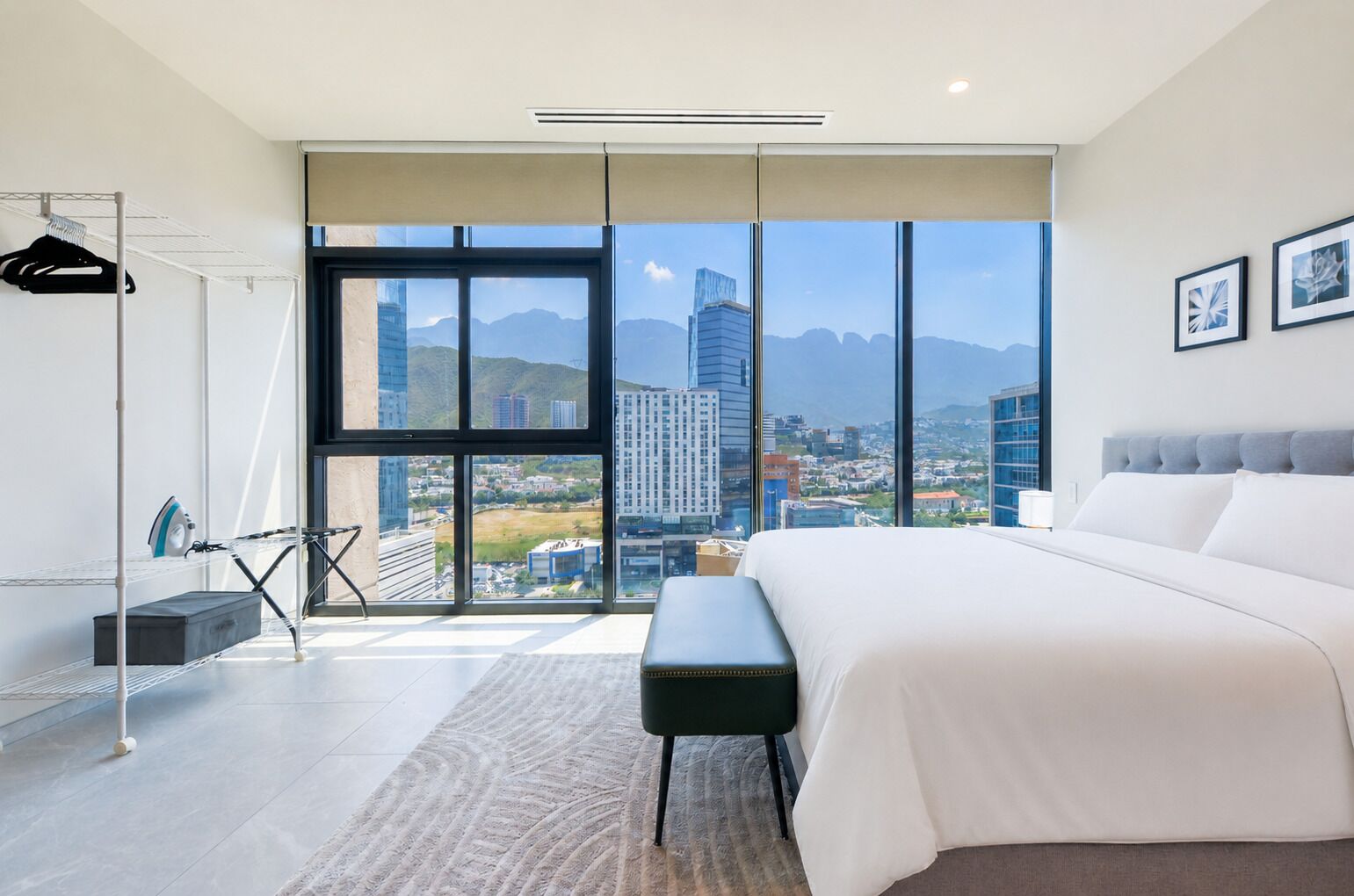 A modern bedroom with a large bed, a bench, a drying rack, and a view of a cityscape through large windows.