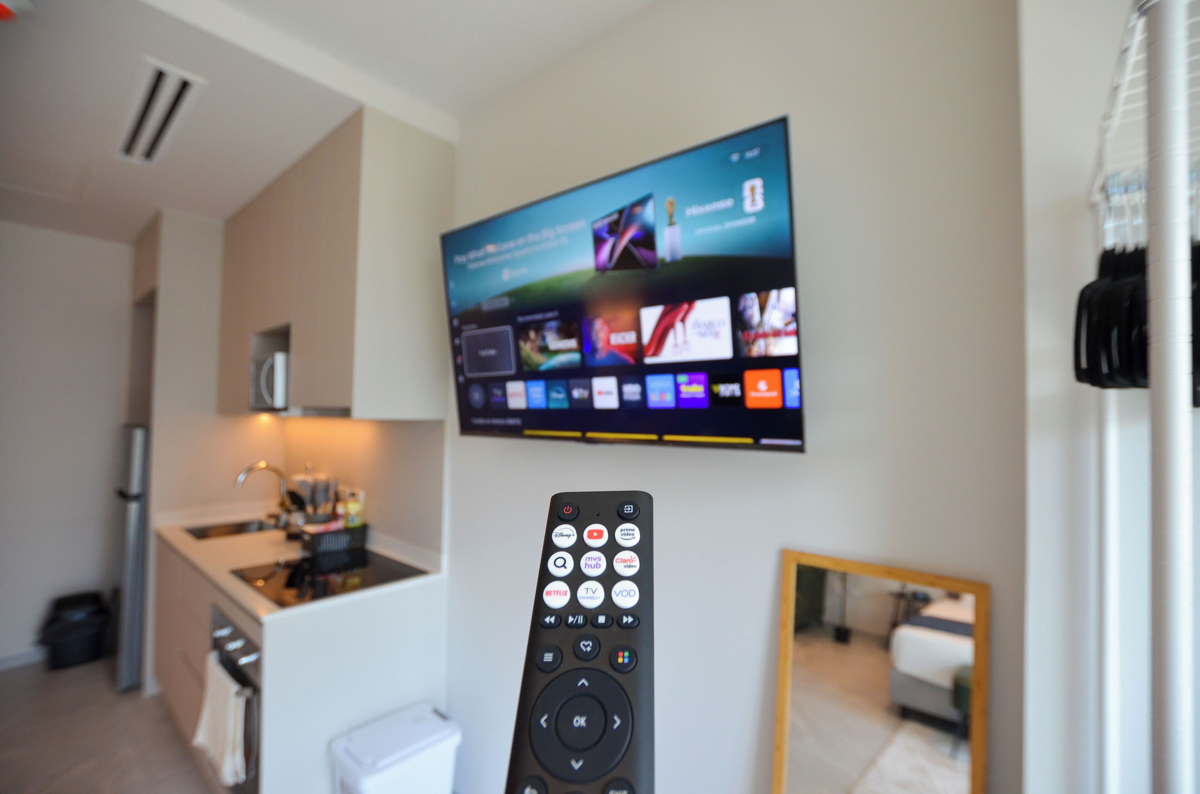 Una sala de estar moderna con un televisor de pantalla plana montado en la pared, un control remoto y una cocina al fondo.