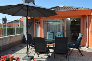 An outdoor patio with a table and chairs, a large umbrella, and a view of the adjacent building. - Cozy bungalow near the beach (Julianadorp)
