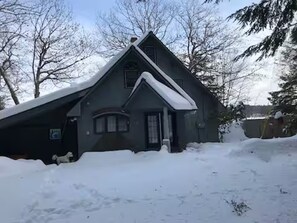 Uma casa com telhado íngreme coberto de neve, rodeada por árvores e um quintal.