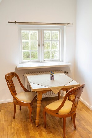 Habitación en esquina con una mesa y dos sillas, un radiador debajo de una ventana y vistas a árboles desde el exterior.