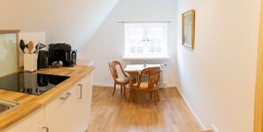 Una cocina pequeña con una placa de inducción negra, sillas de madera, una mesa blanca y una ventana con vistas a árboles.